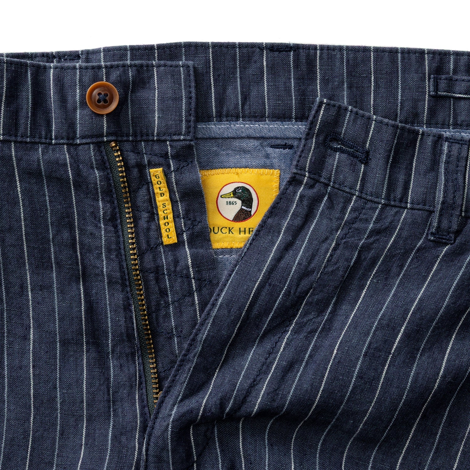 Close-up of Duck Head Gold School Linen Chino Shorts in blue pinstripe, featuring a YKK metal zipper, antique brass button, and partially open to show yellow inner label and detailed stitching.