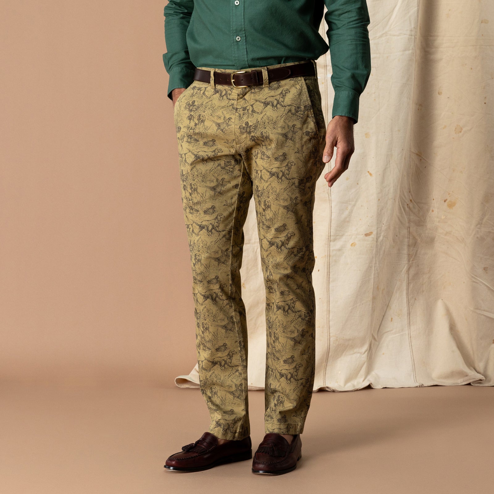 Classic Fit Gold School Chino Wheatfield Print tan pants featuring a repeating illustration of dogs in a field, worn with a dark brown leather belt, green button-down shirt, and brown loafers.