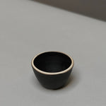 The Stoneware Smudge Bowl by Incausa is a small, black hand-thrown bowl with a light rim, displayed on a smooth light grey surface—ideal for use as a smudge bowl.