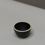 The Stoneware Smudge Bowl by Incausa is a small, black hand-thrown bowl with a light rim, displayed on a smooth light grey surface—ideal for use as a smudge bowl.