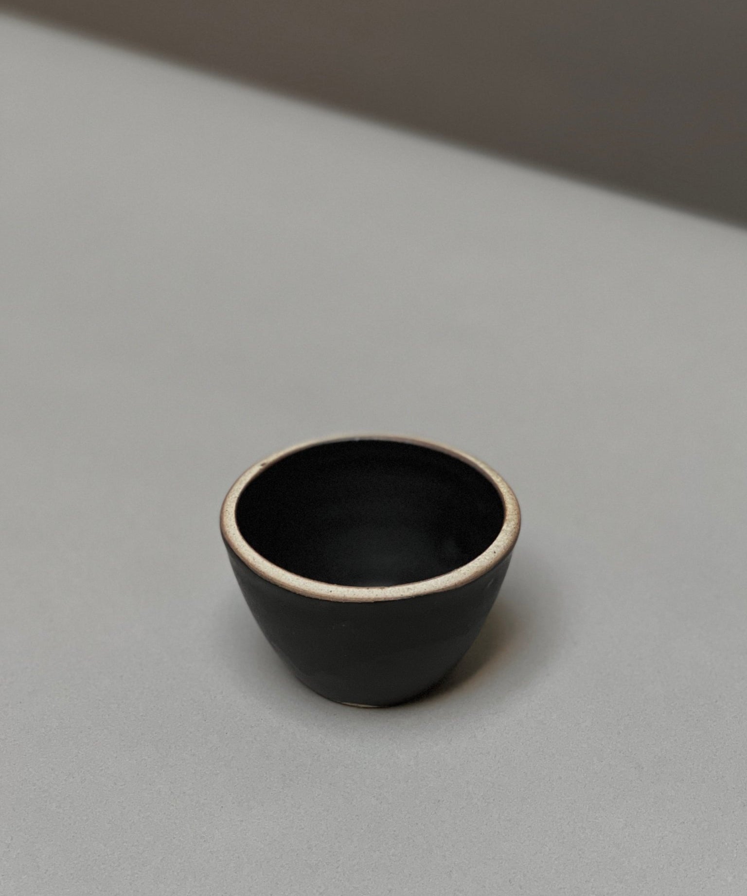 The Stoneware Smudge Bowl by Incausa is a small, black hand-thrown bowl with a light rim, displayed on a smooth light grey surface—ideal for use as a smudge bowl.