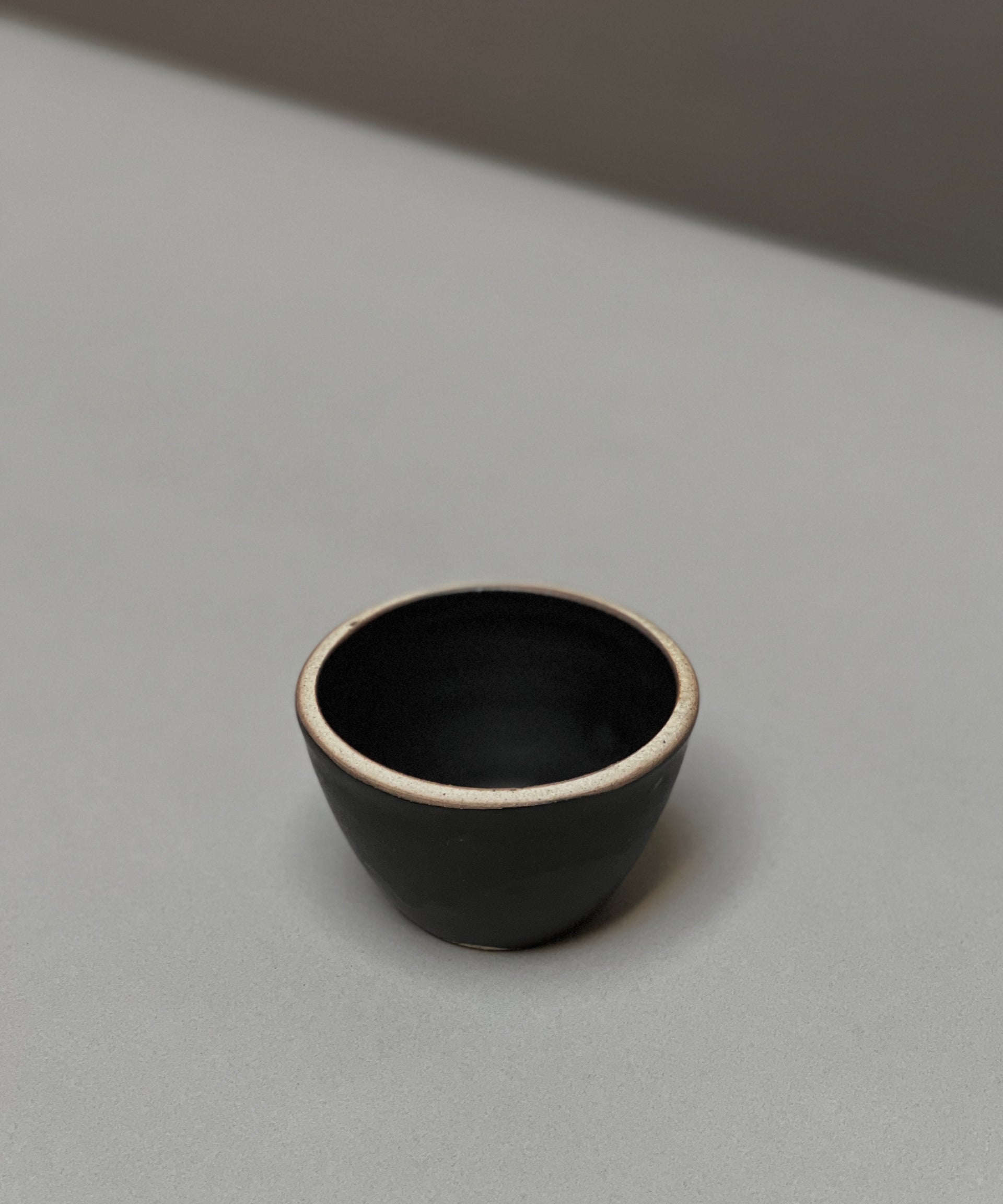The Stoneware Smudge Bowl by Incausa is a small, black hand-thrown bowl with a light rim, displayed on a smooth light grey surface—ideal for use as a smudge bowl.