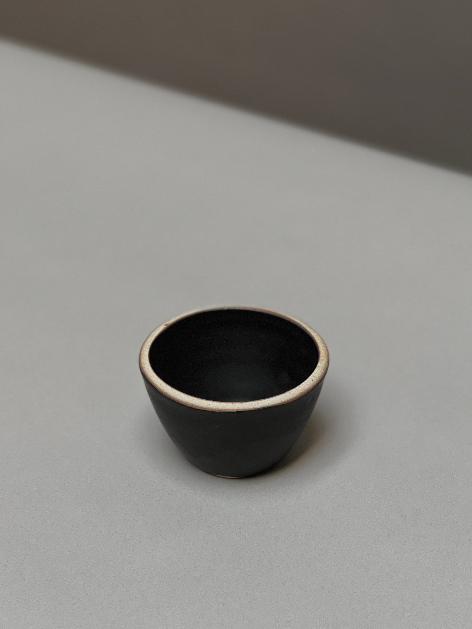 The Stoneware Smudge Bowl by Incausa is a small, black hand-thrown bowl with a light rim, displayed on a smooth light grey surface—ideal for use as a smudge bowl.
