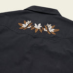 Back view of the Howler Brothers Crosscut Deluxe shirt in dark navy, showcasing white and brown floral embroidery across the upper back western yokes, expertly crafted by Howler Brothers from stretch chambray for comfort and mobility.