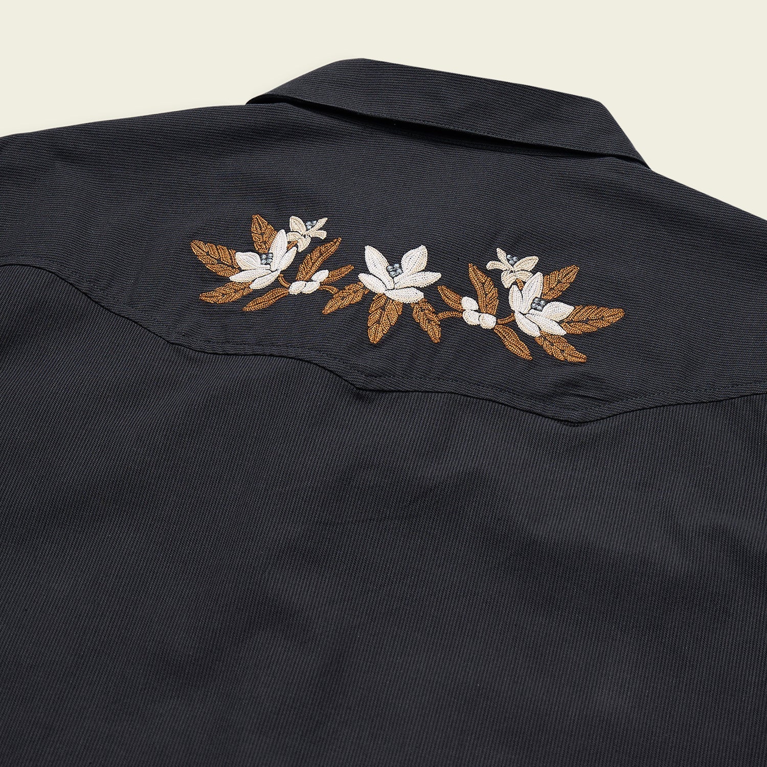 Back view of the Howler Brothers Crosscut Deluxe shirt in dark navy, showcasing white and brown floral embroidery across the upper back western yokes, expertly crafted by Howler Brothers from stretch chambray for comfort and mobility.