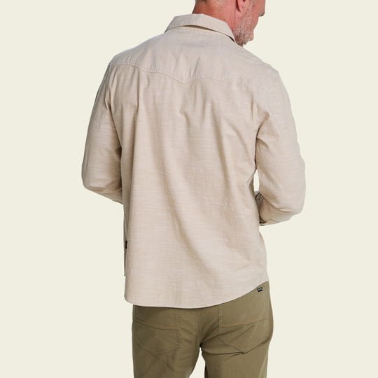 Man facing away, wearing a light beige Howler Brothers Crosscut Deluxe long-sleeve shirt and olive green pants against a plain light background—a relaxed look from the Howler Brothers collection.