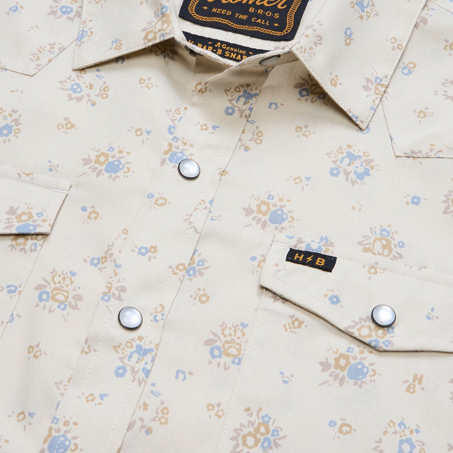 Close-up of the Howler Brothers H Bar B Snapshirt in cream, featuring a floral pattern, two front pockets with pearl snaps, and a small “H.B” tag on the right pocket flap—hallmarks of Howler Brothers’ signature style.