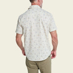 A man is seen from the back wearing the Howler Brothers H Bar B Snapshirt—a short-sleeve, light-colored button-up with a subtle floral pattern—paired with olive green pants.