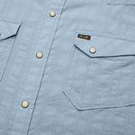 Close-up of a light blue Howler Brothers H Bar B Snapshirt featuring vertical stitching, pearl snaps, and two flap chest pockets—one adorned with a small black and gold “H/B” tag. Designed by Howler Brothers for quick-drying comfort and everyday style.