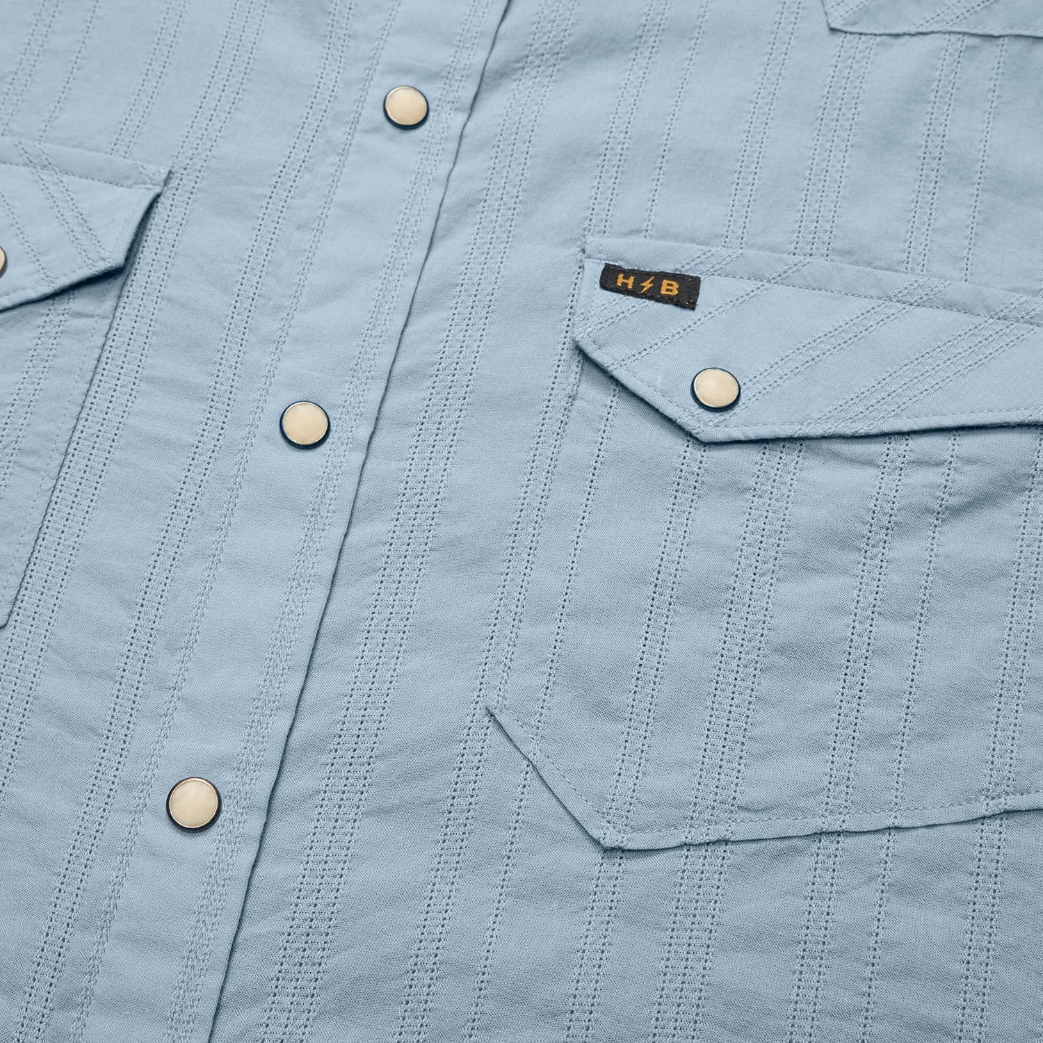 Close-up of a light blue Howler Brothers H Bar B Snapshirt featuring vertical stitching, pearl snaps, and two flap chest pockets—one adorned with a small black and gold “H/B” tag. Designed by Howler Brothers for quick-drying comfort and everyday style.