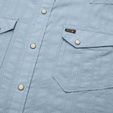 Close-up of a light blue Howler Brothers H Bar B Snapshirt featuring vertical stitching, pearl snaps, and two flap chest pockets—one adorned with a small black and gold “H/B” tag. Designed by Howler Brothers for quick-drying comfort and everyday style.