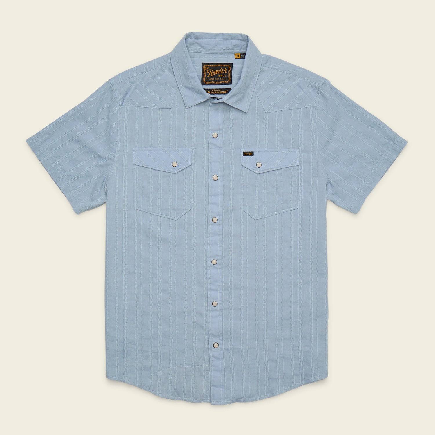 The Howler Brothers H Bar B Snapshirt is a light blue, short-sleeve button-up featuring two chest pockets, subtle vertical stripes, and a branded tag on the left pocket. Designed with classic pearl snaps for easy fastening, this shirt offers quick-drying comfort all day long.