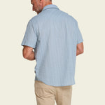 A man is shown from behind wearing the Howler Brothers H Bar B Snapshirt in light blue and beige pants, standing against a plain light background.