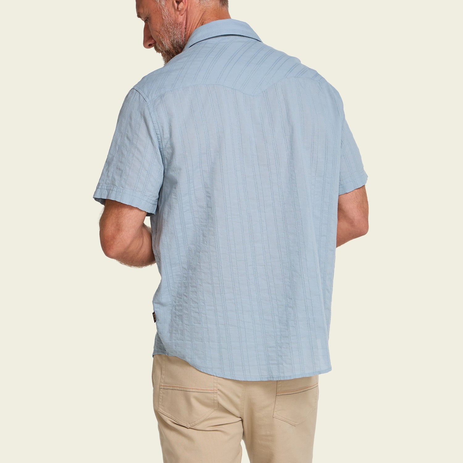 A man is shown from behind wearing the Howler Brothers H Bar B Snapshirt in light blue and beige pants, standing against a plain light background.