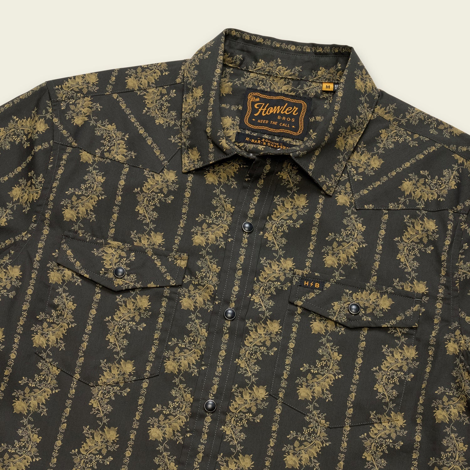 A dark button-up technical utility shirt with a yellow floral stripe pattern, featuring two chest pockets with flaps, pearl snaps for added style, and a “Howler Brothers” label on the inside collar.