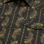 Close-up of a Howler Brothers Grampa’s Curtains black technical utility shirt featuring gold floral patterns, a buttoned front with pearl snaps, two chest pockets with flaps, and a small “H|B” tag above the left pocket.