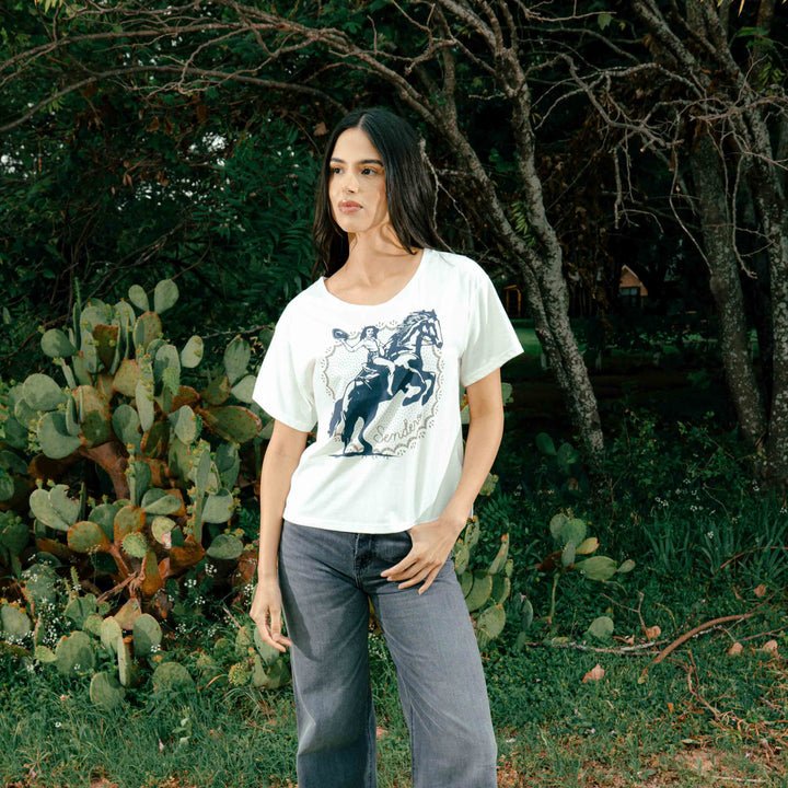 A woman stands outdoors near cacti, wearing the Palomino Basic Tee by Sendero Provisions Co.—a relaxed, roomy white graphic T-shirt crafted from a soft cotton polyester blend—paired with gray jeans, with trees and greenery in the background.