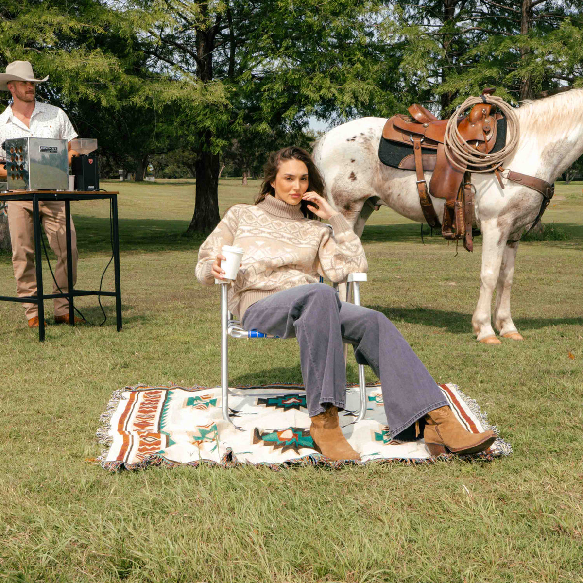 A woman in a southwest-inspired Shiloh Sweater and dark jeans sits on a folding chair on a patterned blanket, with a saddled horse and a man making coffee nearby in a grassy field.