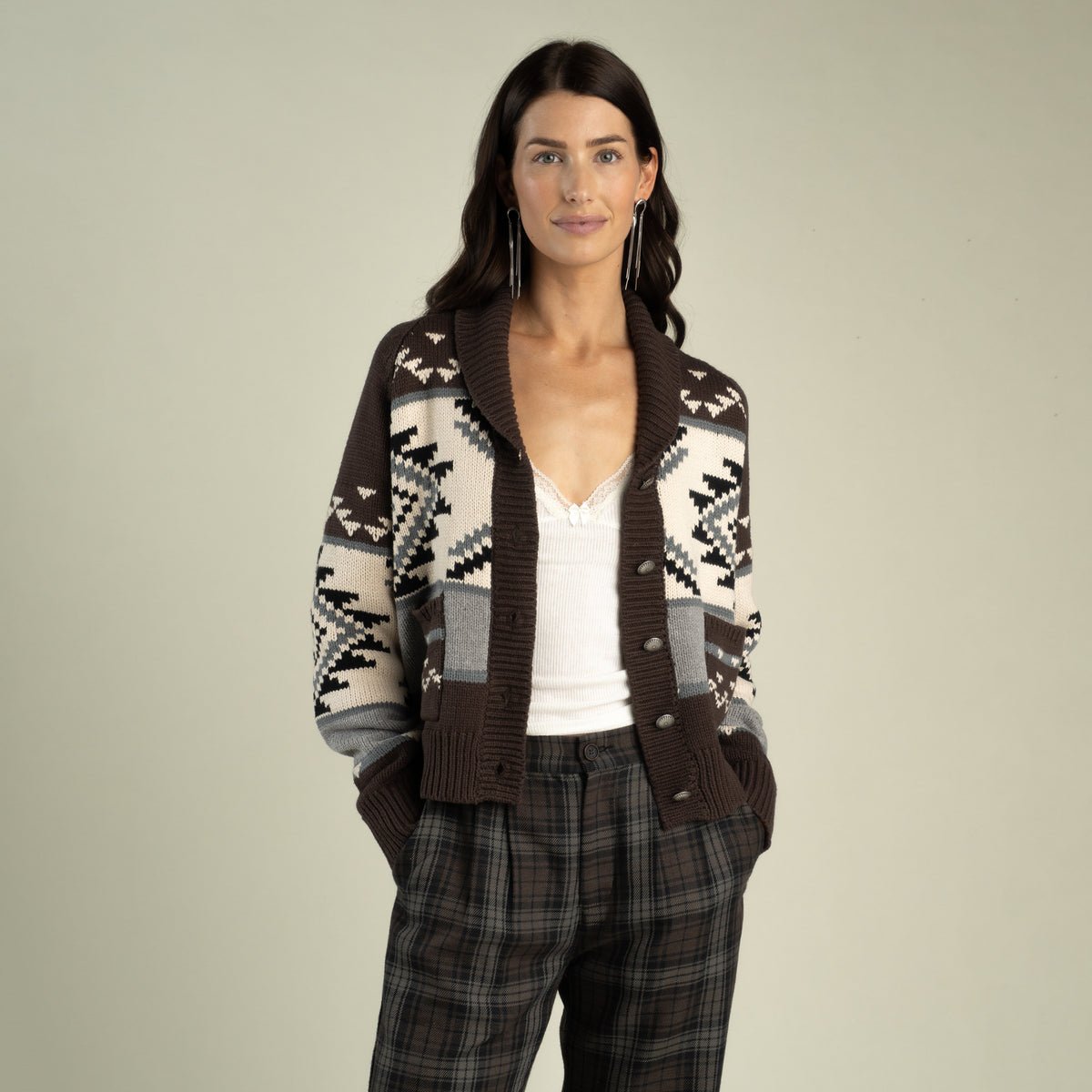 A woman wears a brown, cream, and grey Cimarron Cardigan by Sendero Provisions Co. with a shawl collar over a white camisole and plaid trousers.