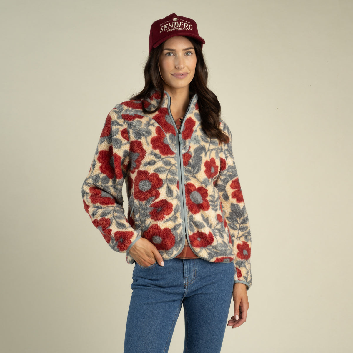A woman wears a Sedona Fleece by Sendero Provisions Co. in a red and grey southwest print, paired with blue jeans and a burgundy baseball cap.