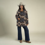 A person wearing a grey cowboy hat, a brown and blue Santa Fe Jacket by Sendero Provisions Co., dark wash wide-leg jeans, and brown boots.