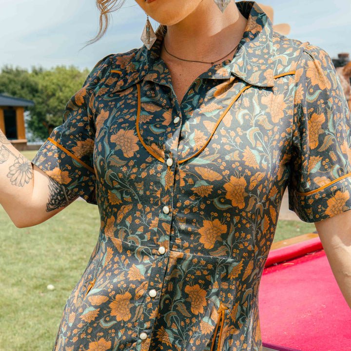 A woman wearing the La Dahlia Dress by Sendero Provisions Co.—a short-sleeve, floral print western dress in orange and green hues—stands outdoors beside a red pool table.