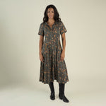 A woman stands and smiles against a plain light background, wearing the La Dahlia Dress by Sendero Provisions Co.—a dark floral print western dress with short sleeves and pearl snaps—paired with black boots.