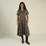 A woman stands and smiles against a plain light background, wearing the La Dahlia Dress by Sendero Provisions Co.—a dark floral print western dress with short sleeves and pearl snaps—paired with black boots.