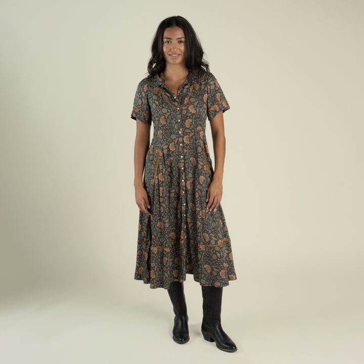 A woman stands and smiles against a plain light background, wearing the La Dahlia Dress by Sendero Provisions Co.—a dark floral print western dress with short sleeves and pearl snaps—paired with black boots.