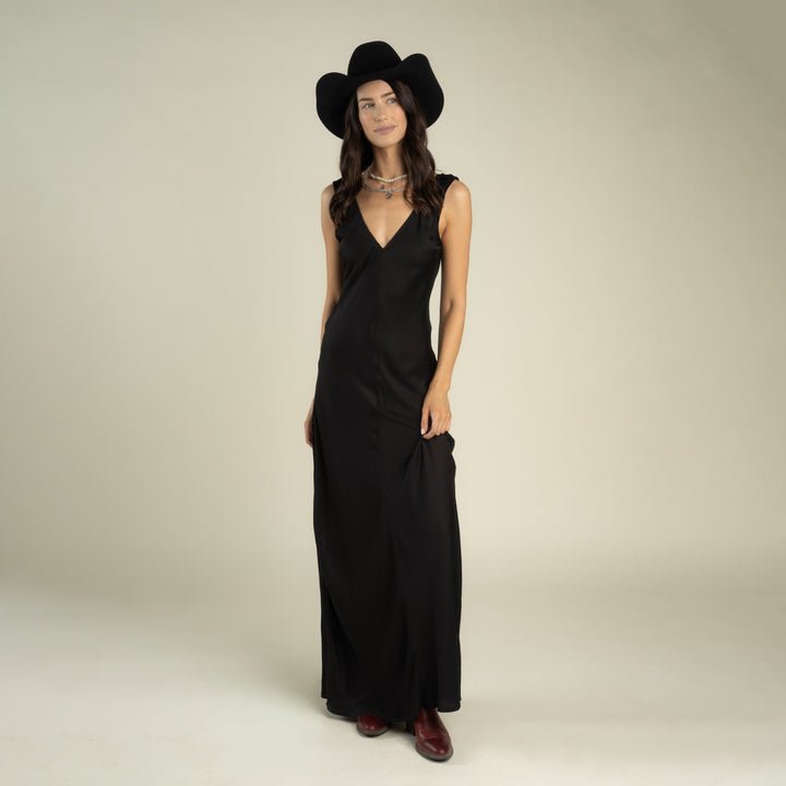 A woman wearing the Sendero Provisions Co. Romy Slip Dress, paired with a black wide-brimmed hat and brown boots, stands against a plain light background.