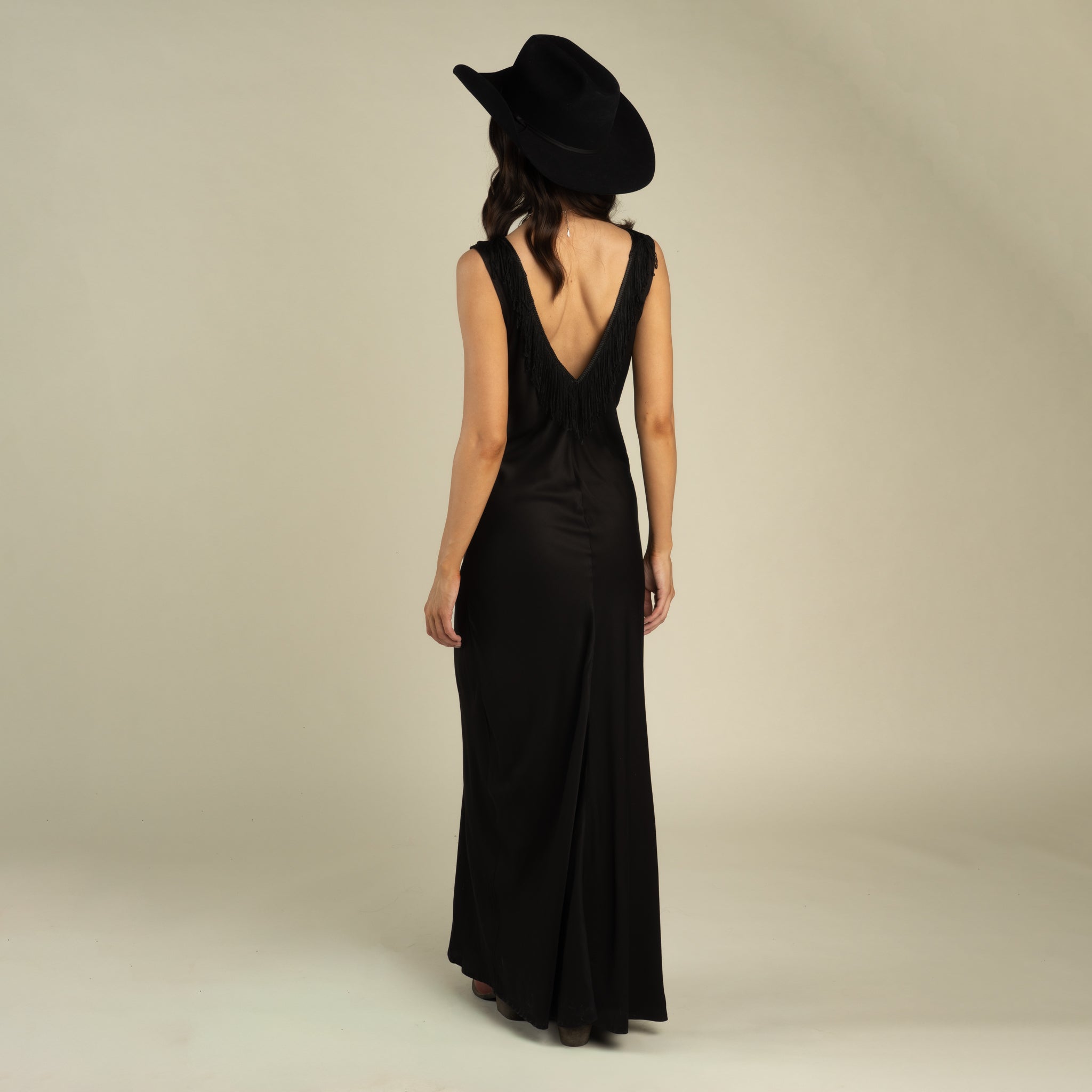 A woman stands facing away, wearing the Romy Slip Dress by Sendero Provisions Co.—a black viscose maxi dress with a graceful bias fit—paired with a wide-brimmed black hat, set against a plain light background.