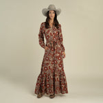 A woman wearing a gray wide-brim hat, the Miradora Dress by Sendero Provisions Co.—a long-sleeve Desert Floral linen maxi dress with a self belt at the waist—and brown boots, standing against a plain light background.