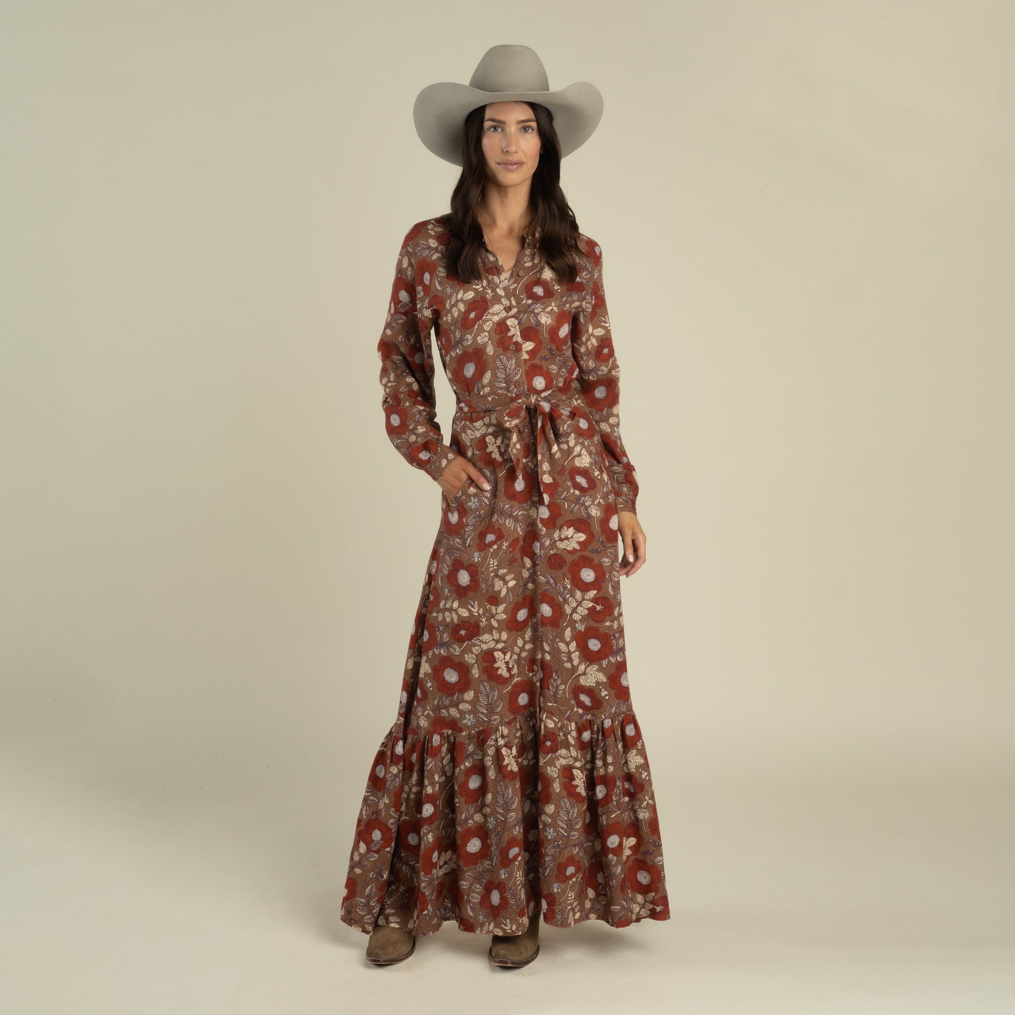 A woman wearing a gray wide-brim hat, the Miradora Dress by Sendero Provisions Co.—a long-sleeve Desert Floral linen maxi dress with a self belt at the waist—and brown boots, standing against a plain light background.