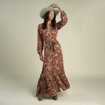 A woman stands in a studio, holding the brim of her wide-brimmed hat and looking to the side, dressed in the Miradora Dress by Sendero Provisions Co.—a long, Desert Floral linen maxi dress with a self belt.