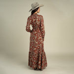 A woman wearing a wide-brimmed hat and the Sendero Provisions Co. Miradora Dress—a long, Desert Floral linen maxi dress with a self belt—stands facing away against a plain background.