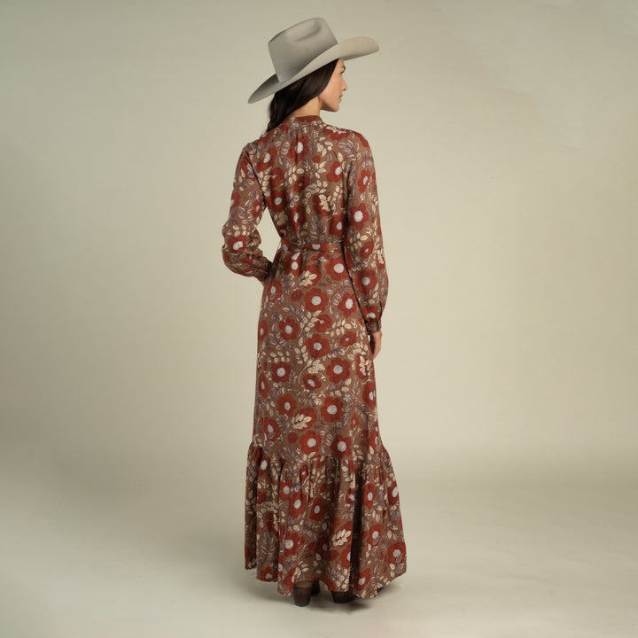 A woman wearing a wide-brimmed hat and the Sendero Provisions Co. Miradora Dress—a long, Desert Floral linen maxi dress with a self belt—stands facing away against a plain background.