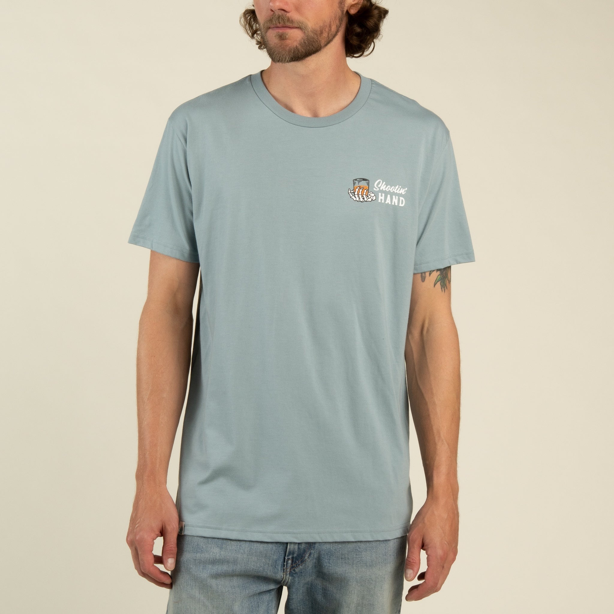 A man wearing a light blue Sendero Provisions Co. Shootin’ Hand t-shirt, crafted from ultra soft cotton with a small “Steady Hand” logo and tiger graphic on the left chest in soft touch screen print, paired with light blue jeans, stands against a plain background.