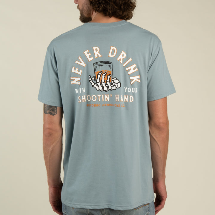 Wearing the Sendero Provisions Co. Shootin’ Hand t-shirt by Sendero Provisions Co.—a light blue, ultra-soft cotton shirt featuring a soft touch screen print graphic of skeleton hands holding a glass and the western proverb: “Never drink with your shootin’ hand.”.