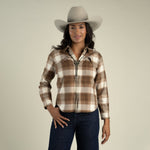 A woman wearing a brown and white Sendero Women’s Durango Flannel - Pearlsnap, dark jeans, and a light-colored cowboy hat with a bolo tie.