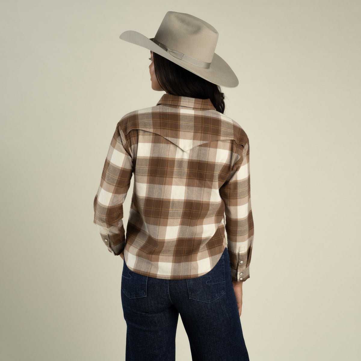 A woman viewed from behind wearing a Sendero Women’s Durango Flannel - Pearlsnap in brown and white brushed cotton with a western-style back yoke, dark jeans, and a light gray cowboy hat.