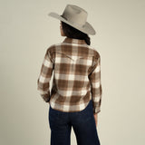 A woman viewed from behind wearing a Sendero Women’s Durango Flannel - Pearlsnap in brown and white brushed cotton with a western-style back yoke, dark jeans, and a light gray cowboy hat.