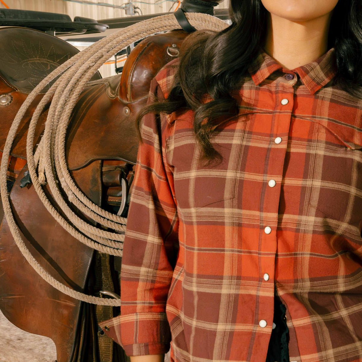 A woman wearing a rust and brown plaid Sendero Women’s Durango Flannel - Pearlsnap, standing next to a western saddle with a rope.