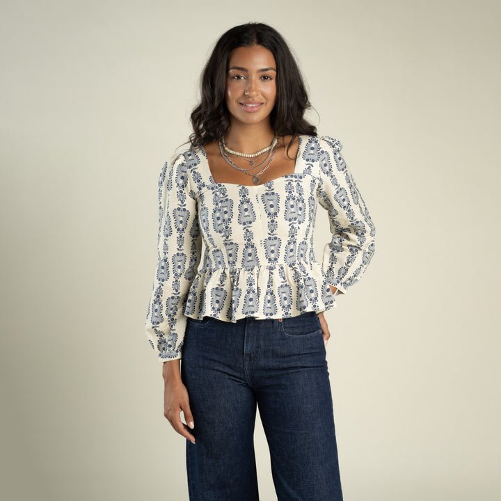 A woman stands against a plain beige background, smiling slightly with one hand on her hip, wearing the Ada Blouse by Sendero Provisions Co.—a patterned long-sleeve blouse with a flattering contoured fit—paired with dark blue jeans.