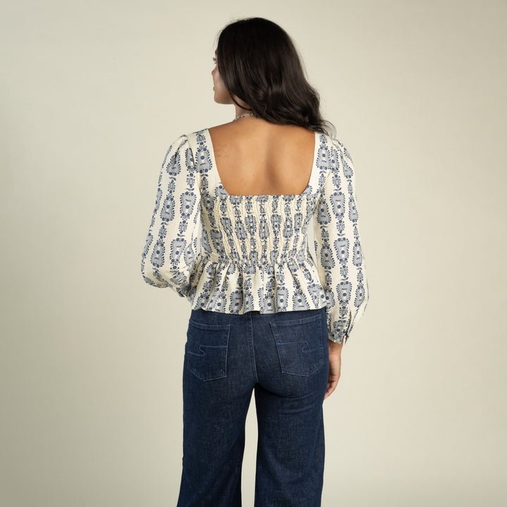 A woman faces away, wearing the Ada Blouse by Sendero Provisions Co.—a patterned long-sleeve blouse featuring a square neckline and smocked back for a contoured fit—paired with dark blue jeans and standing against a plain light background.
