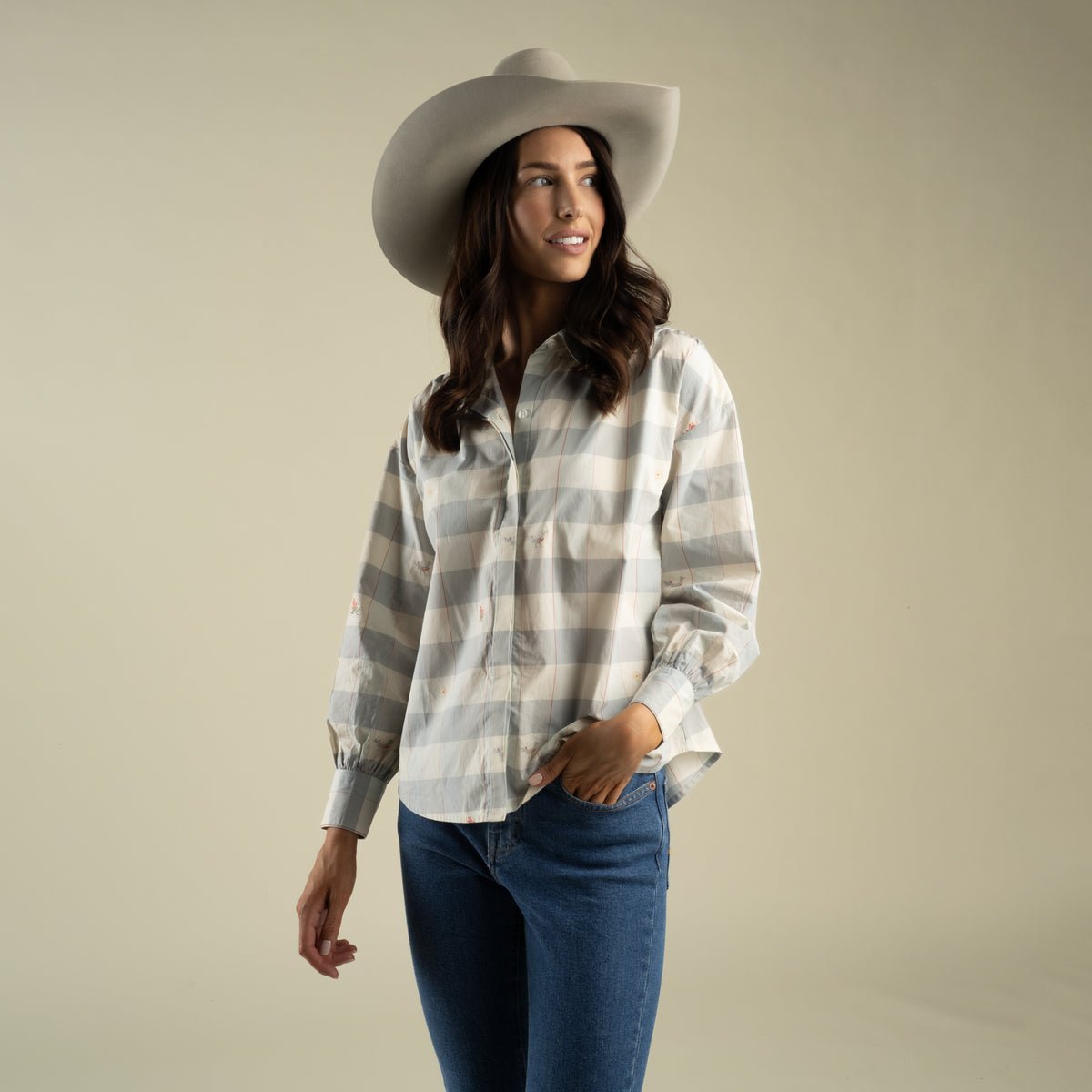 A woman wearing a tan cowboy hat and the Sendero Helena Button Down, a grey and white paisley check shirt with small floral embroidery, tucked into blue jeans.