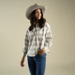 A woman wearing a tan cowboy hat and the Sendero Helena Button Down, a grey and white paisley check shirt with small floral embroidery, tucked into blue jeans.