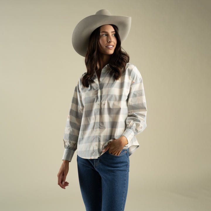 Wearing the Helena Button Down by Sendero Provisions Co.—a gray and white plaid shirt crafted from 100% cotton—paired with blue jeans and a wide-brimmed hat, a woman stands against a plain beige background, looking off to the side with one hand in her pocket.