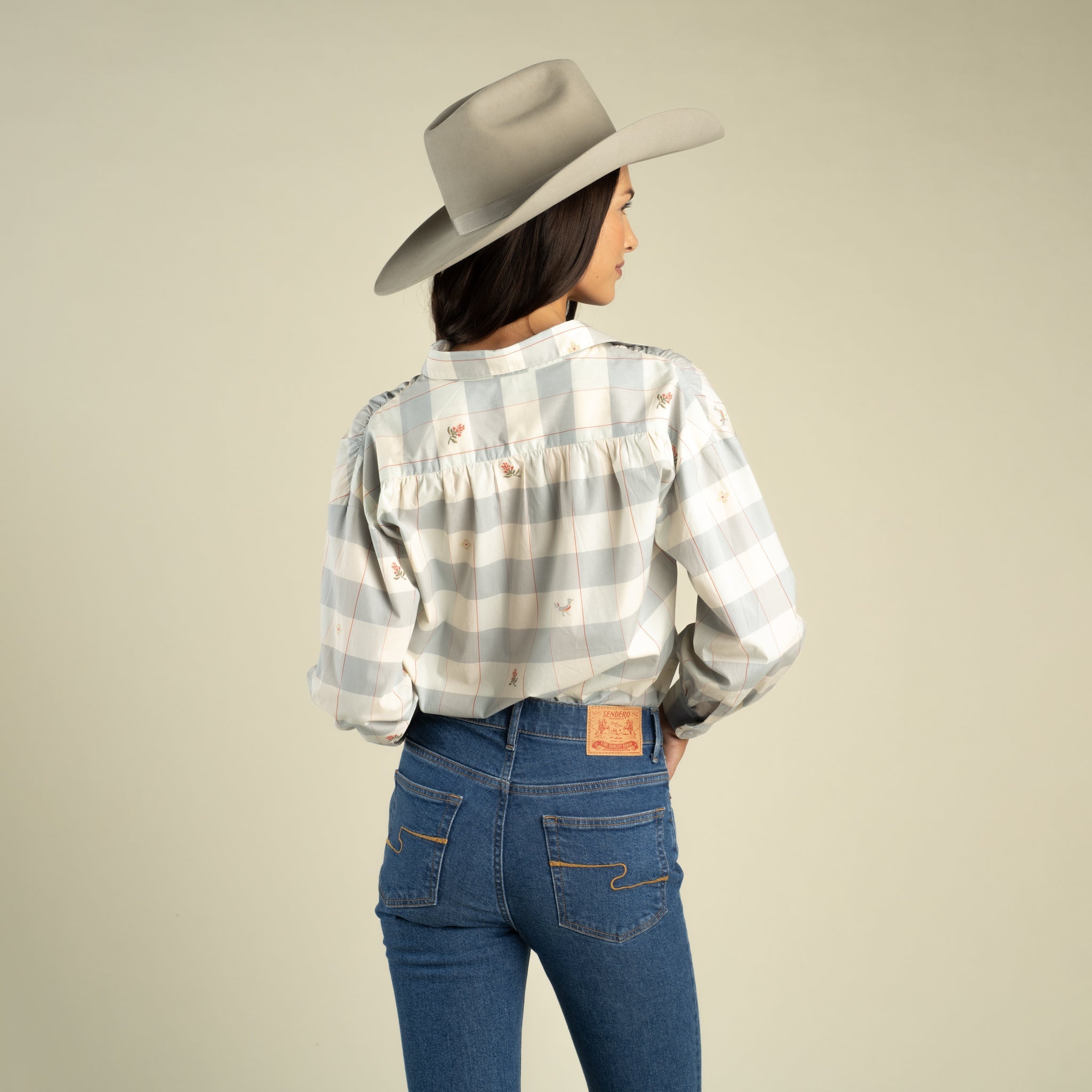 A woman in the Sendero Provisions Co. Helena Button Down stands facing away, highlighting the relaxed fit and effortless style of her plaid shirt paired with blue jeans, all set against a plain background.