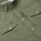 Close-up of The Grazer | Poncho, an olive green work shirt by Poncho, featuring front flap pockets with silver snap buttons and a small black "Poncho" label on the left pocket.