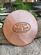 Add a touch of style to any space with the Ballad of the Bird Dog 5 Pack Leather Coaster, featuring a round leather patch embossed with “Ballad of the Bird Dog, Brenham, TX.” Each coaster rests on a textured surface and is accented with leaves in the background—ideal as handmade coasters or stylish desk accessories for any setting.
